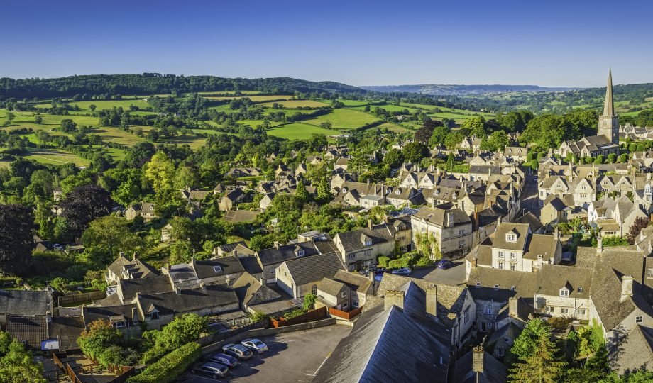Aerial panorama over idyllic country village cottages green summer fields Clear blue summer skies and vibrant green patchwork fields above the iconic Cotswold village of Painswick, with its honey coloured limestone cottages and historic church spire. Representative of nature and biodiversity.